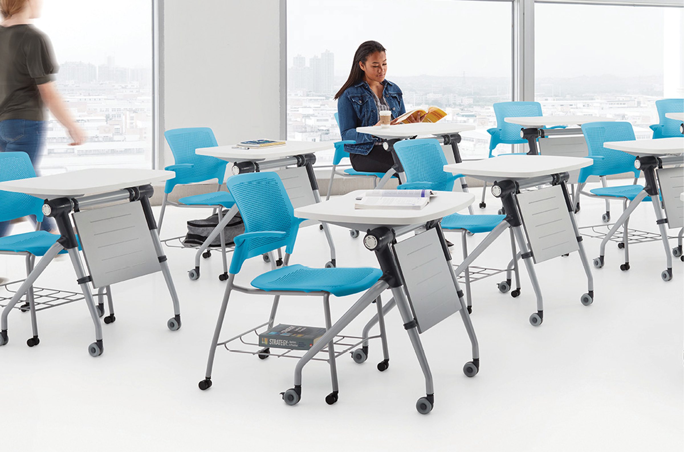 Woman reading a book and drinking coffee in room with two rows of classroom style desks with chairs